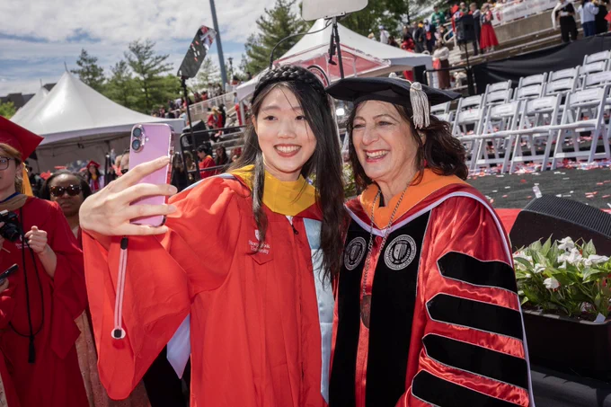 A Stony Brook graduate is taking a selfie with me. We are Both are smiling and standing amidst the graduation ceremony setting with chairs and people in the background. 