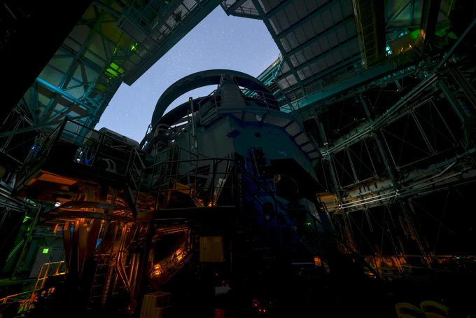 Night view inside an observatory: a massive teal telescope structure and amber lighting, framed by the open dome and a star-filled sky overhead.