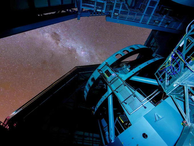 Wide-angle view inside an observatory: a large teal-blue telescope mount and cables frame an open roof, revealing a star-filled sky with the glowing band of the Milky Way.