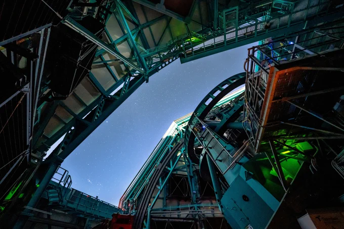 Upward view inside a large observatory dome at night with teal steel beams, platforms, and  telescope mount under an opening to a starry sky.