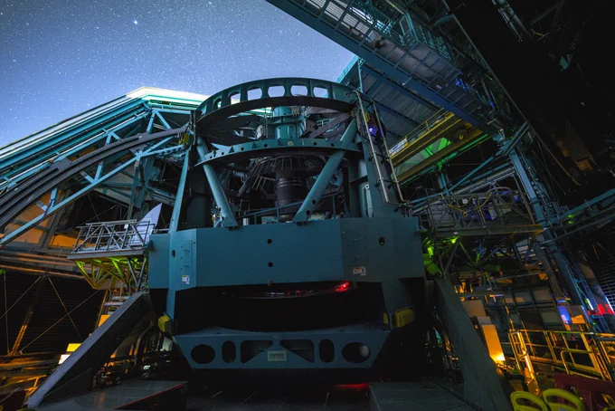 Night view inside an open observatory dome with a massive teal telescope mount beneath a starry sky, surrounded by metal walkways and green and orange LEDs.