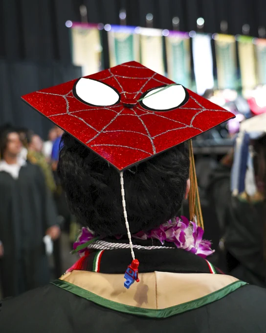 A graduate is wearing a decorated cap featuring a Spider-Man design with a web pattern and eyes. The tassel is hanging down, and a small Spider-Man keychain is attached.