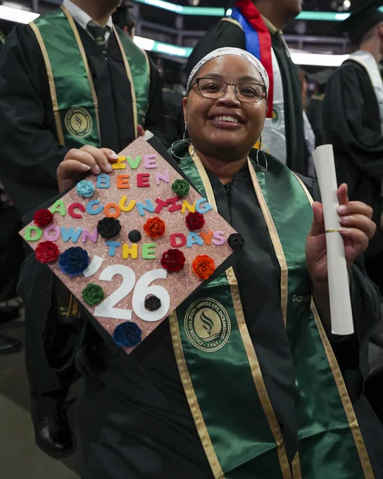 A graduate holds up a decorated graduation cap with colorful flowers and the words "I've been accounting down the days: 26." The individual also holds a rolled diploma.