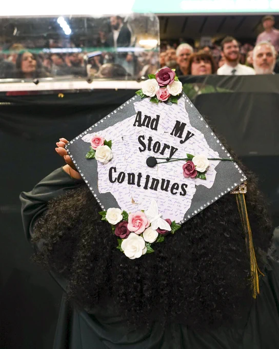 A graduate's cap decorated with artificial flowers and the words "And My Story Continues." It appears during a ceremony, with people seated and watching in the background.