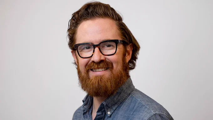 Man with curly brown hair and beard wearing a blue button-up shirt against a plain white background.
