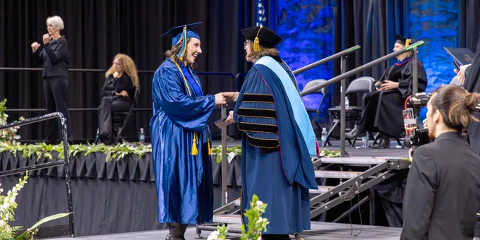 A person in a blue graduation gown shakes hands another person on stage.
