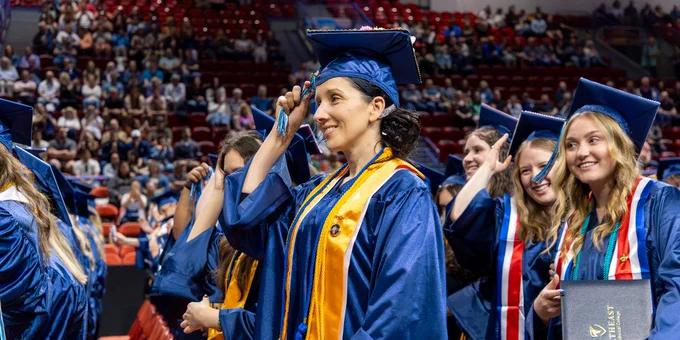Graduates in blue caps and gowns stand in a crowded auditorium. 