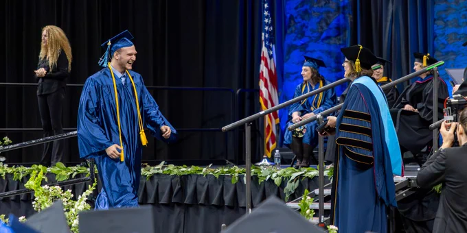 A graduate in a blue cap and gown walks across a stage to receive a diploma during a commencement ceremony.