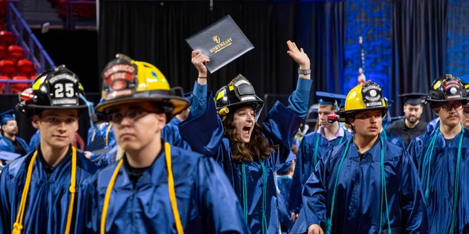 A group of people in blue graduation gowns wearing firefighter helmets.