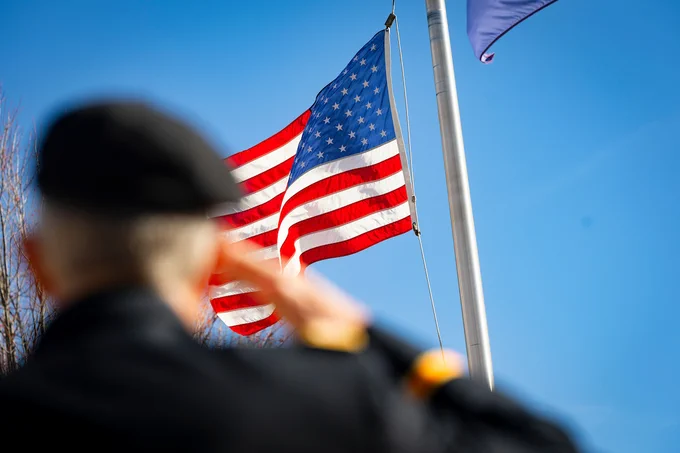 A person in uniform salutes the United States flag as it waves on a flagpole against a clear blue sky.
