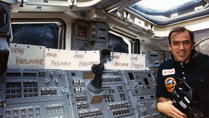 John Fabian, STS-51G mission specialist, posing with "Deliver" and "Pickup" signs lining space shuttle Discovery's aft flight deck. The signs are in reference to the satellite deploys and retrieval that were achieved during the June 1985 mission. (NASA)