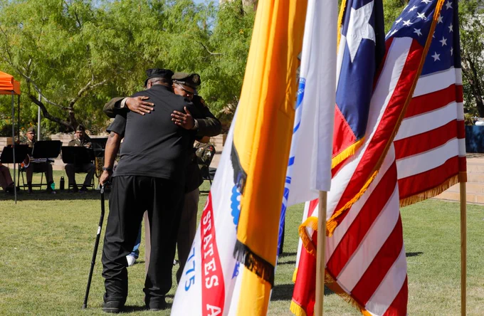 Two individuals embrace near colorful flags, including the Texas and United States flags, at the ROTC commissioning ceremony at UTEP's Centennial Plaza. A group of musicians is visible in the background under a canopy.
