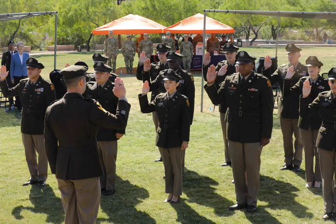 A group of uniformed military personnel stands outdoors on a grassy area, raising their right hands in UTEP's ROTC Commissioning ceremony. In the background, people are gathered under orange canopies. UTEP's Centennial Plaza is visible.