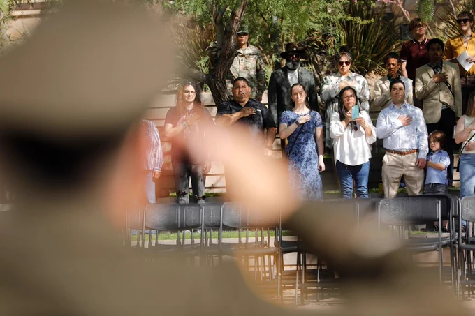 A group of individuals stands with their hands over their hearts during UTEP's ROTC Commissioning ceremony. A person in military attire is visible in the foreground, with a focus on their salute. Some attendees stand beside empty chairs, and others are gathered near greenery in the background.