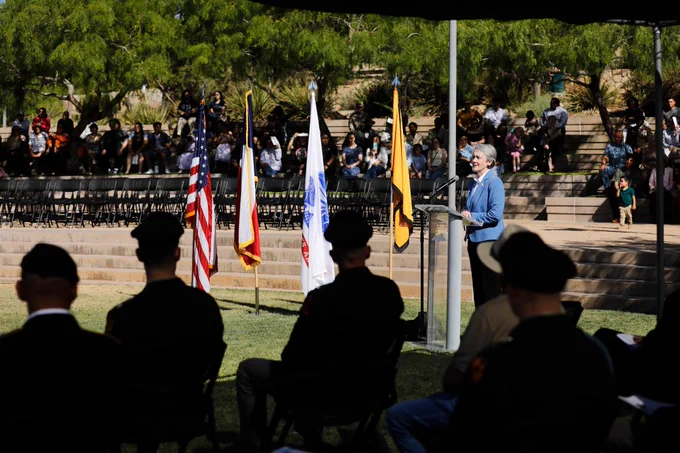 UTEP President Heather Wilson speaks at a podium in UTEP's ROTC Commissioning Ceremony with the U.S., Texas, and other flags displayed nearby. Attendees, some in military attire, are seated in rows on tiered steps, and the setting features green trees in the background.