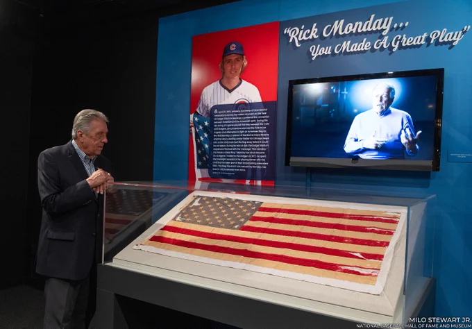 Rick Monday admires a Museum exhibit dedicated to the United States flag he rescued on April 25, 1976. A worn flag is enclosed in a case accompanied by wall art, a monitor and the quote "Rick Monday... you made a great play." (Milo Stewart Jr./National Baseball Hall of Fame and Museum)