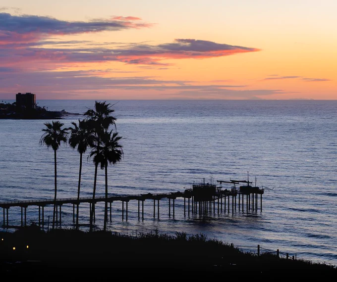 A scenic view of Scripps pier extending into the ocean during sunset, with silhouettes of palm trees in the foreground.