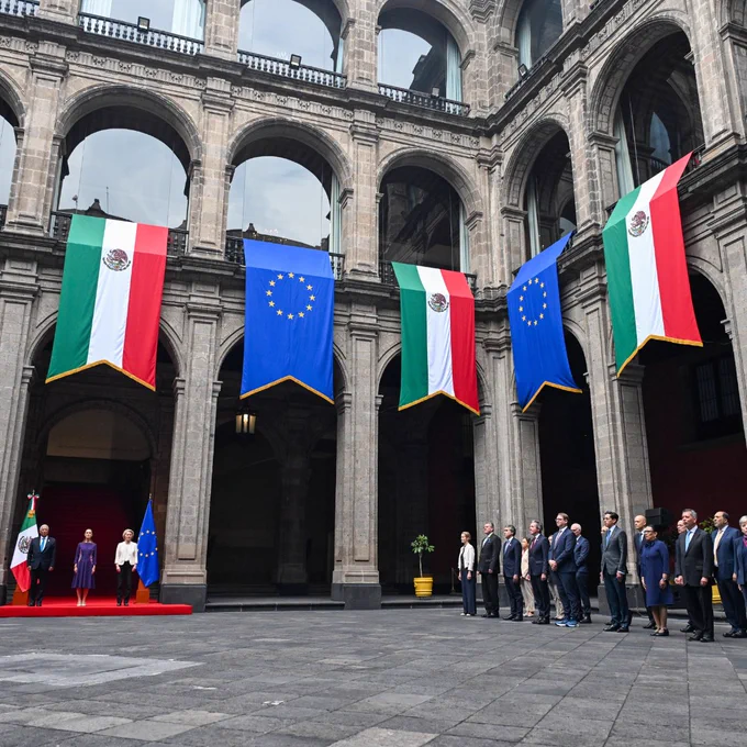 A formal ceremony in a stone courtyard, with large Mexican and European Union flags hanging from the arches above two facing lines of officials from Mexico and the EU standing on opposite sides of the square.