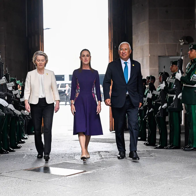 Ursula von der Leyen, Claudia Sheinbaum and António Costa walk side by side through a grand doorway, passing between two lines of uniformed guards holding rifles in a ceremonial honour guard.