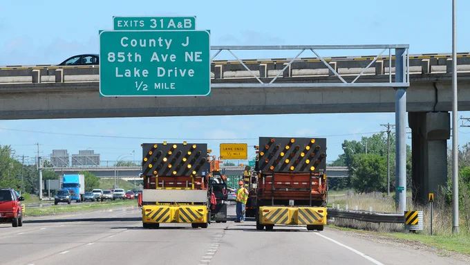 MnDOT maintenance vehicles with striped barriers working on a highway under the County J, 85th Avenue Northeast, and Lake Drive exit sign. Workers are positioned near the vehicles, and a bridge is visible in the background.