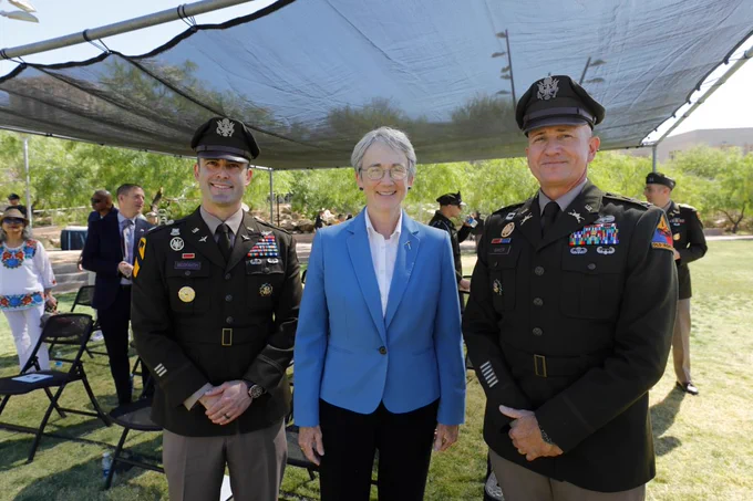 Two individuals in military uniforms and UTEP President Heather Wilson, wearing a blue blazer, standing outdoors at an ROTC commissioning event. They are under a canopy with trees in the background and additional people seated nearby.
