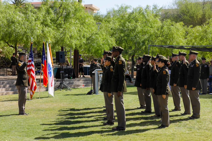 A group of 14 UTEP ROTC cadets standing in formation during an outdoor ceremony. Several flags, including the American flag, are displayed on the left. Trees and a small audience are visible in the background.