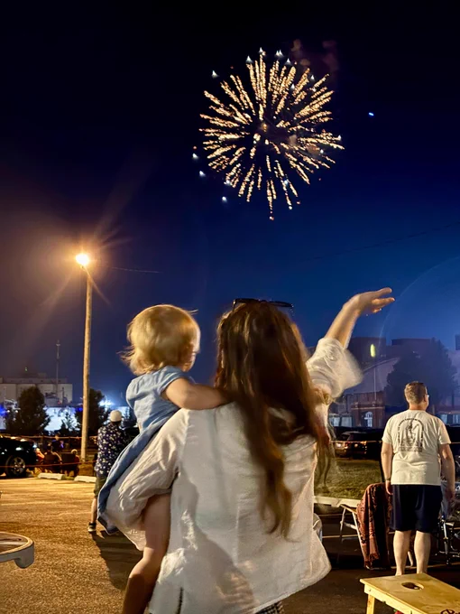 A person is holding a child while pointing at colorful fireworks in the night sky. There are buildings and parked cars in the background, with streetlights illuminating the area.