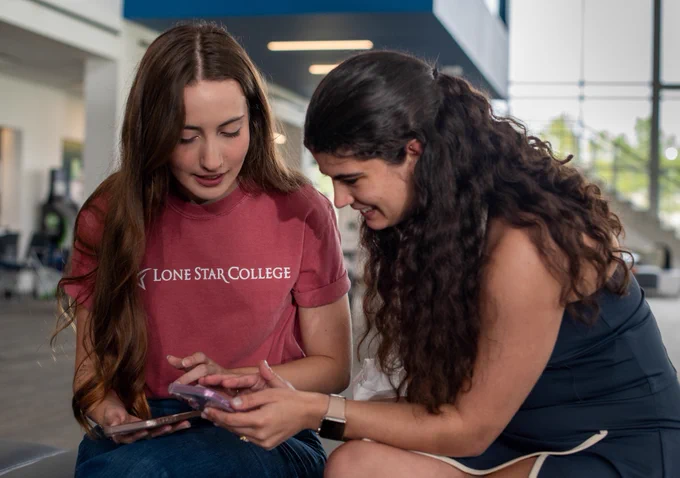 Two individuals sitting together, both looking at a smartphone. One is wearing a Lone Star College T-shirt. They appear to be indoors, in a bright and spacious area.