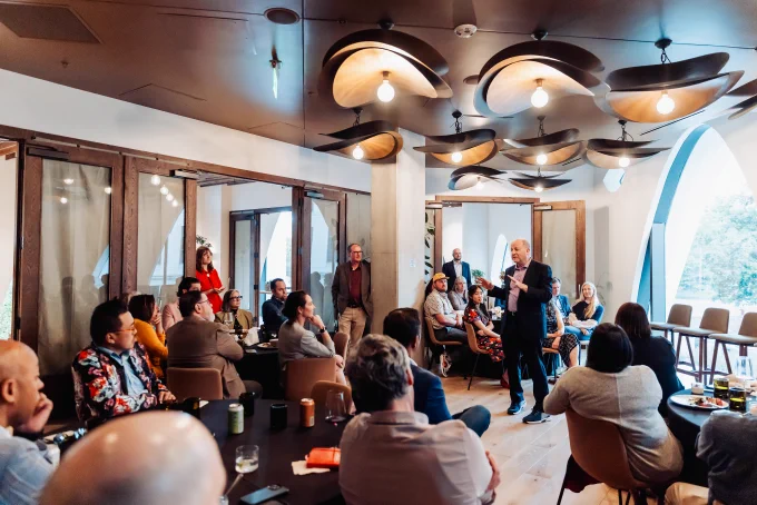 Governor Jared Polis addresses a diverse group seated in a modern room with wooden accents and unique ceiling lights during a professional event.