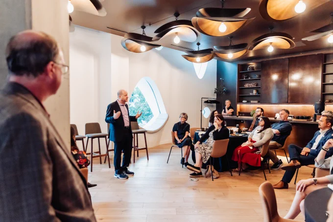 Governor Jared Polis addresses a diverse group seated in a modern room with wooden accents and unique ceiling lights during a professional event.