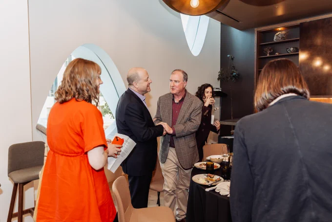 Governor Jared Polis addresses a diverse group seated in a modern room with wooden accents and unique ceiling lights during a professional event.