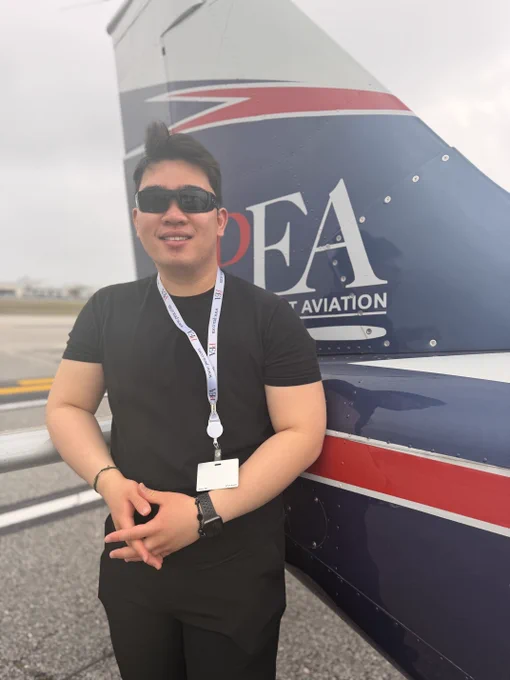 A person wearing sunglasses stands beside a small aircraft with "L3Harris Flight Academy" branding on the tail fin. The individual is smiling and holding a lanyard with a badge. The background shows an overcast sky above an airport runway.