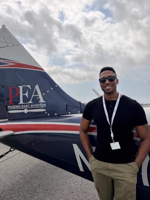 The image shows a person standing beside the tail of an aircraft with "Phoenix East Aviation" and "PEA" visible on it. The individual is smiling and wearing sunglasses, a black shirt, khaki pants, and a lanyard with an ID badge. The sky is partly cloudy.