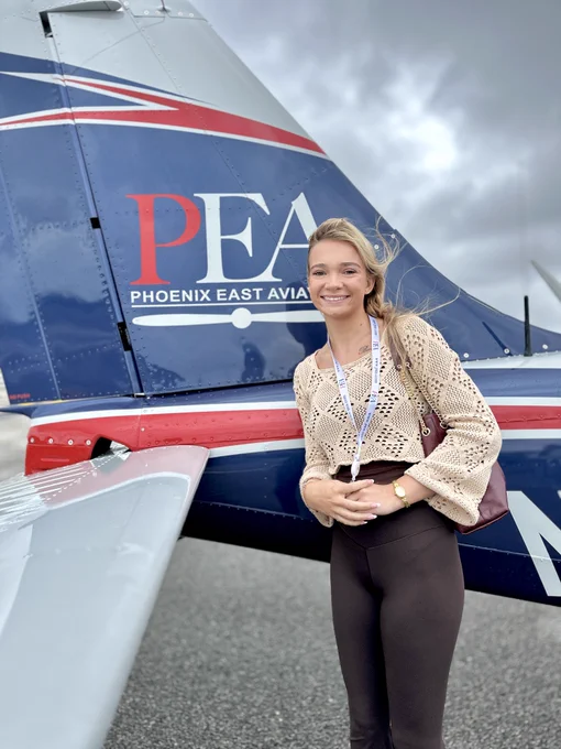 A person stands in front of a plane with "Phoenix East Aviation" and the initials "PEA" visible on the aircraft's tail. The individual is smiling and wearing a crocheted sweater, black pants, and a lanyard. The sky is overcast.