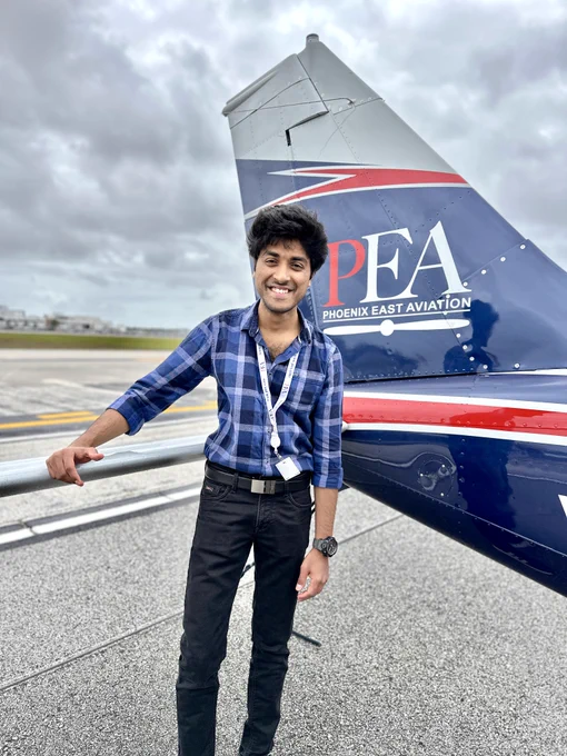 Smiling person standing on an airport tarmac beside the tail of a small airplane with "Phoenix East Aviation" written on it. They are wearing a blue checkered shirt and a lanyard with an ID badge.