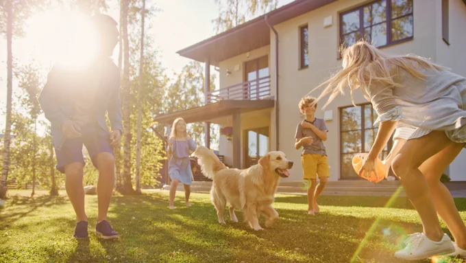 A group of people playing with a golden retriever on a sunlit lawn in front of a modern two-story house with large windows.