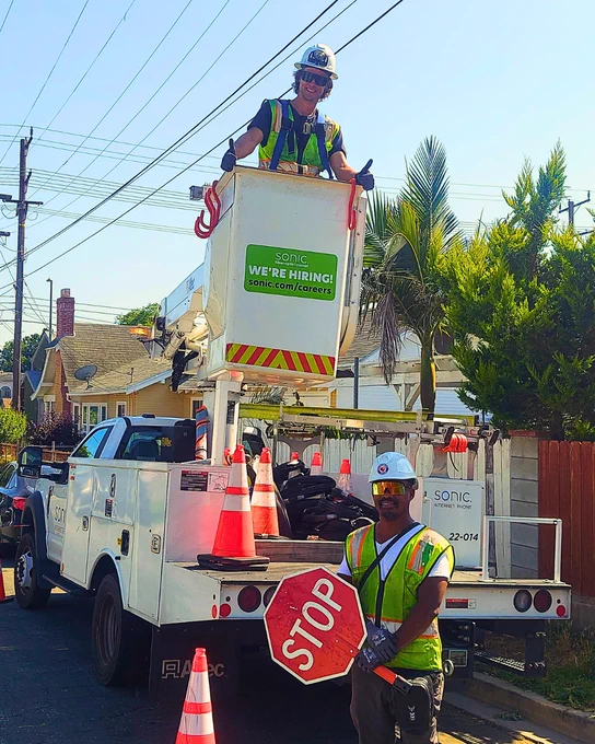 Two Sonic Field Service team members in Southern California, one smiling and throwing two thumbs up from within the bucket on a Sonic bucket truck and the other smiling and standing in front of the back of the truck, holding a Stop sign. 