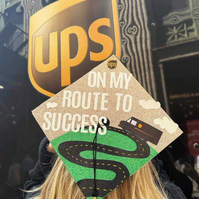 A graduate wearing a graduation cap with a road design and the words \"ON MY ROUTE TO SUCCESS\" in front of a UPS truck.