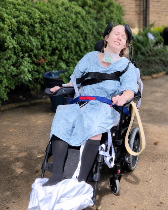 A young woman in a wheelchair, wearing a medical gown and neck brace, appears distressed outdoors among lush greenery.