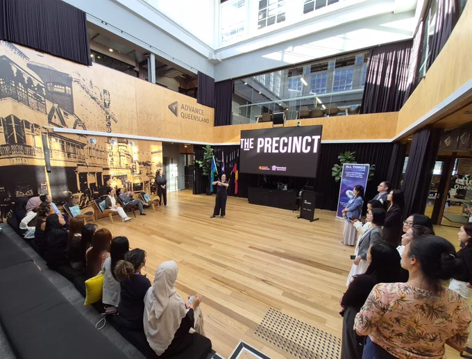 A speaker addressing participants seated in a modern indoor venue with 'THE PRECINCT' displayed on a large screen behind them.