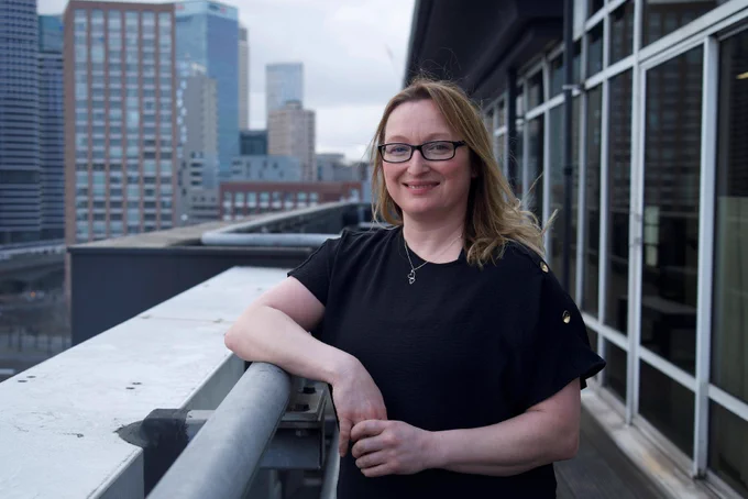 The image shows an individual standing on a balcony with a cityscape backdrop. Tall skyscrapers, including some with glass facades, are visible in the background. The scene appears to be during the day with a cloudy sky.