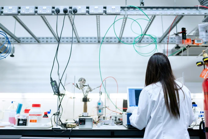 Scientist in a white lab coat working at a cluttered laboratory bench with various scientific instruments and cables overhead.