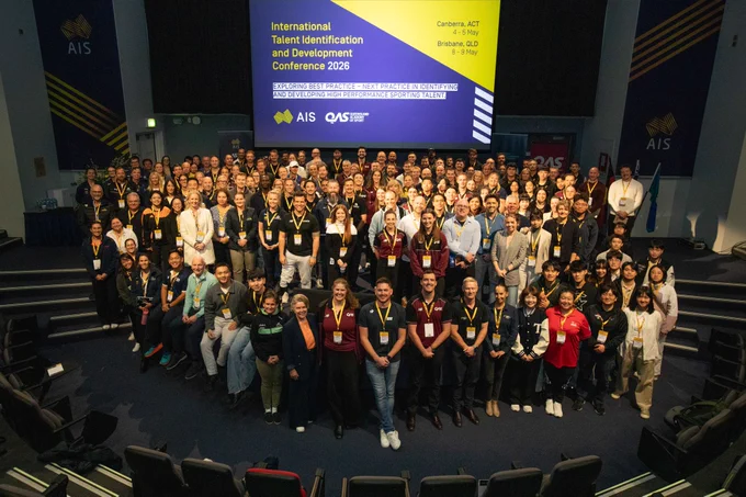 A large group of people is gathered in an auditorium for the International Talent Identification and Development Conference 2026. They are facing the camera, and many are wearing lanyards. A screen displays event details, including locations in Canberra, ACT, and Brisbane, QLD. The AIS and OAIS logos are visible.
