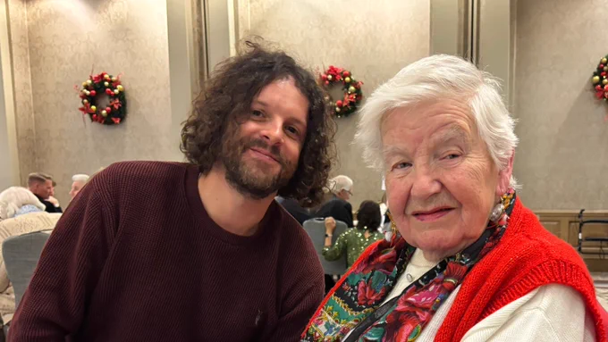 Mike is a man in his 30s with wavy brown hair and facial hair, he is sat down and leaning towards Mary who is also sat down. Mary is in her 80s and has white hair. Both are looking towards the camera and smiling