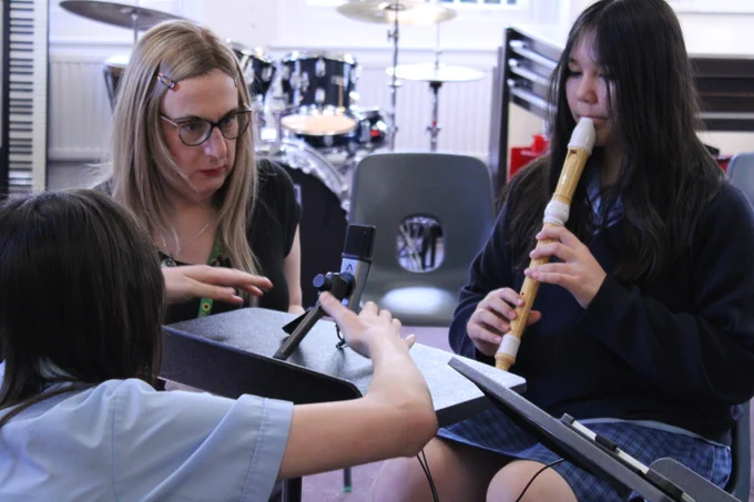 Two students and a tutor in a music classroom, one playing a recorder while another adjusts a microphone on a stand nearby.