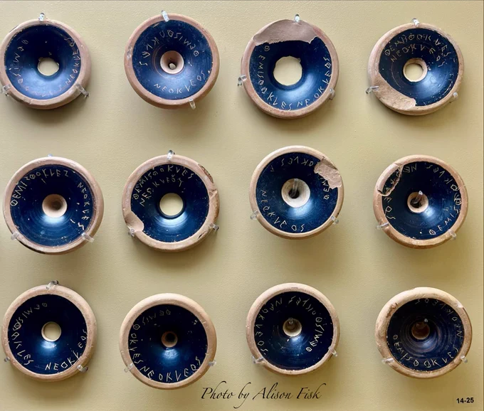 My photo shows twelve black painted circular bases from a type of ancient Greek pottery wine cup known as a ‘kylix’. At a glance they look like a display of chocolate ring doughnuts as they have a central hole, a dark painted ring, and a light clay outer ring. They are arranged in a museum display case in three rows of four, one row above the other. Each piece is inscribed by hand with the name of the famous Athenian general and politician Themistocles. They were pre-prepared for use as ‘ballots’ in a democratic voting process known as an ostracism. Broken pottery sherds like this, used as ballots for voting in an ostracism, are known as ‘ostraca’ (plural) / ‘ostracon’ (singular).

These were found in a trove of pre-prepared ballots in a well on the north slope of the Athenian Acropolis. Seemingly unused, it’s suggested they were pre-prepared to hand out to Athenian voters who couldn’t write. Whilst Themistocles was not ostracised on that occasion, he was exiled a decade later.