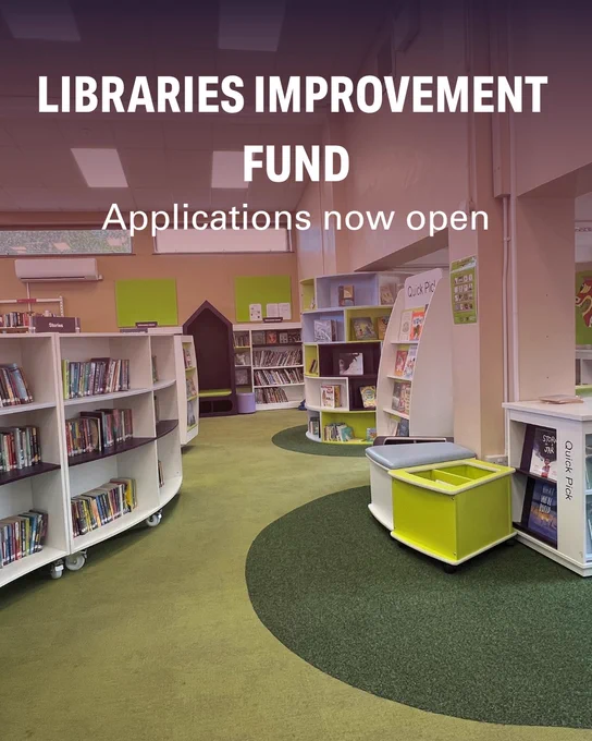 A library interior with bookshelves filled with books. A sign reads, "Libraries Improvement Fund: Applications now open." The carpeting is green with circular patterns.
