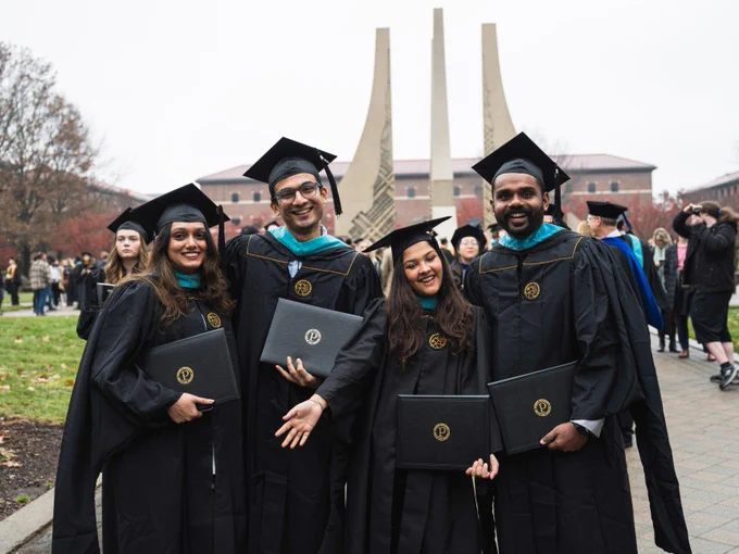 A group of four people wearing graduation caps and gowns stand proudly holding diplomas. They are outdoors, with two tall, narrow structures visible in the background. Other graduates and attendees are seen in the background among fall foliage.