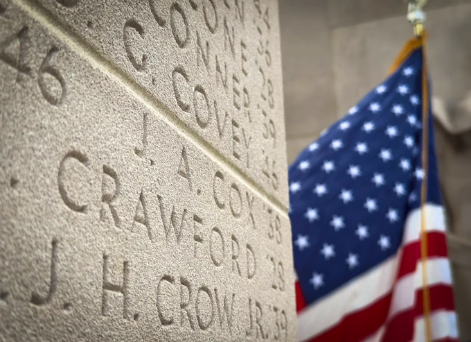 Several names carved on the Pylons war memorial are closely in focus with an American flag positioned behind the names. The wind has unfurled the flag as it moves in the breeze.
