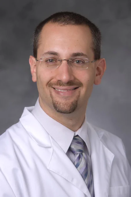 Professional headshot portrait of Mustafa Bashir. He is posing against a gray background and smiling. He is wearing a white lab coat and a multicolored tie.